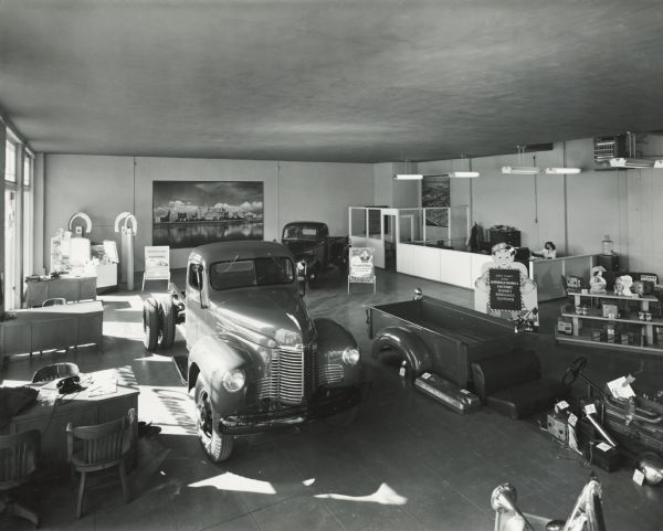 Showroom of the Long Beach branch house featuring International trucks and parts on the showroom floor. There is an enlarged photograph or mural displayed on the back wall. A woman is on the telephone in an office area on the right.