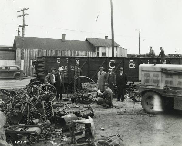 A group looks on as a man uses a cutting torch to reduce scrap iron into readily marketable pieces in the farm scrap collection drive in Barton County. According to the original caption, the scrap drive netted 550 tons in one week. A freight car is being loaded with scrap.