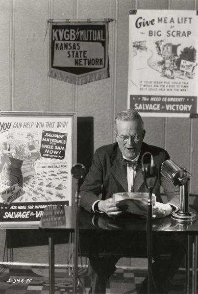 News broadcaster Bon Turner makes an appeal over the radio to farmers to bring their scrap metal into town to help meet the country's need for vital war material. Behind him are posters for "Salvage for Victory."
