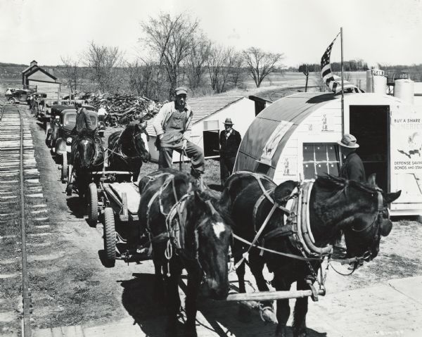 Men with horses, wagons, cars and trucks are parked alongside railroad tracks near a large scrap metal pile. The men are participating in a wartime scrap drive.