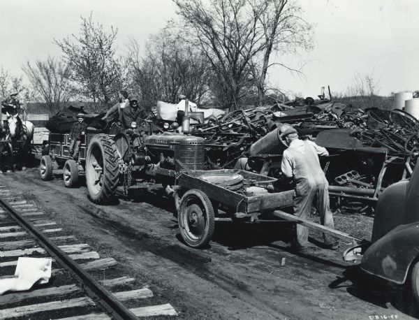 Men and boys use horses, a Farmall H tractor and wagons to haul scrap metal to a pile near railroad tracks. The men are participiting in a wartime scrap drive. The man on the Farmall H is likely Helmuth Strobush. His six year old son Jerry is hanging on the wagon behind the tractor.