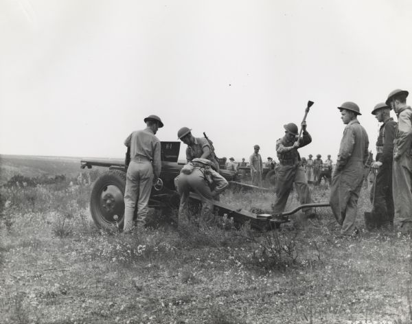 Group of men, some with shovels and axes, setting up a gun on a artillery range. A large group of men in the background are setting up more guns.