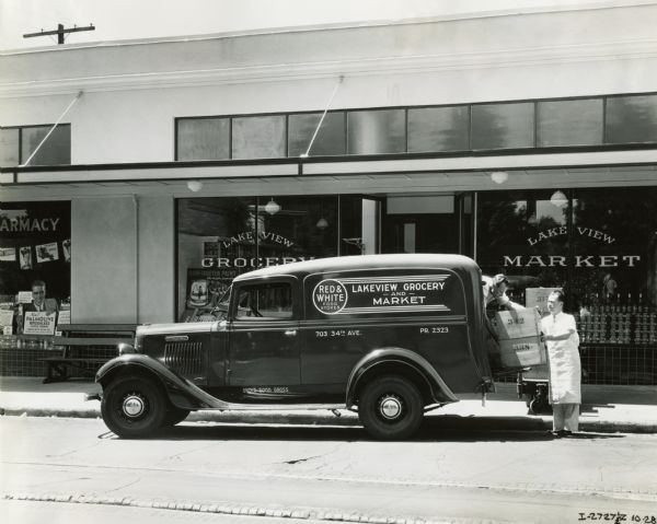 Men load boxes into the back of an International C-1 truck owned by Lakeview Grocery. The truck is parked in front of the Lakeview Grocery storefront. Text on the side of the truck reads: "Red & White Food Stores."