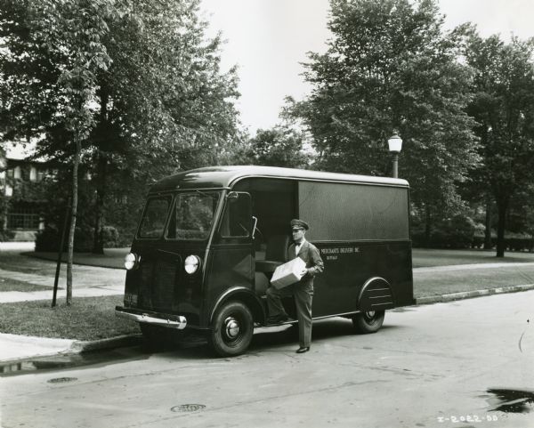 A uniformed delivery man is standing next to an International D-15 truck parked near the curb in a residential neighborhood.