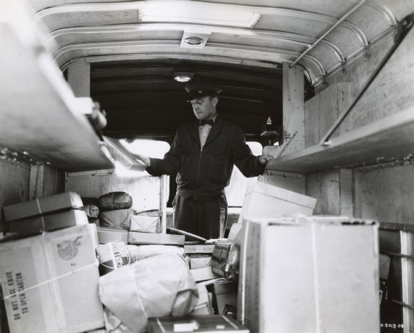 View from rear of the interior of an International D-15 truck. A uniformed man is standing inside among a pile of packages.