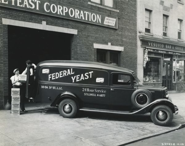 Men loading boxes of yeast into the back of an International truck owned by the Federal Yeast Company. The truck is parked on a sidewalk in front of the garage door of the Federal Yeast Corporation service shop. Next door is the Vesuvio Bar & Grill.
