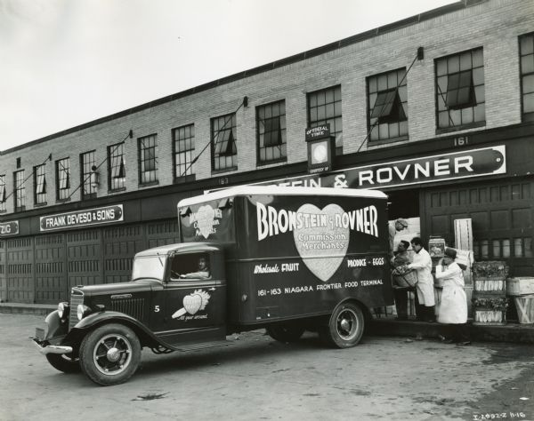 Four men load fruits and vegetables into the back of an International C-30 truck owned by Bronstein & Rovner. The truck is backed up at the loading dock of the Bronstein & Rovner building.