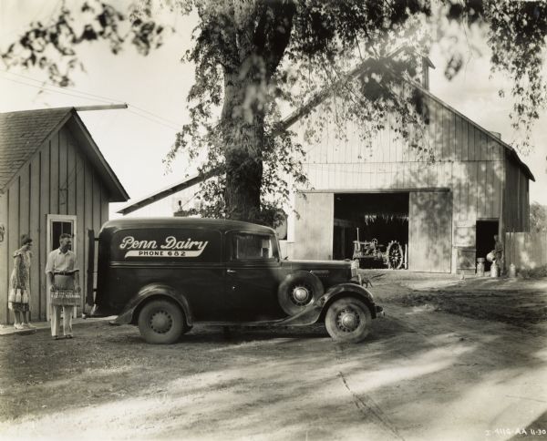 A man and woman are loading crates of milk into an International C-Line truck at Penn Dairy Farm. According to the original caption, "Mr. and Mrs. Johhnson" are "at right"(?). A Farmall Regular(?) tractor is visible through an open barn door in the background.