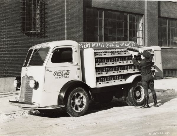A man is loading crates of bottled Coca-Cola into an International C-300 truck parked in the street in front of a large brick building.