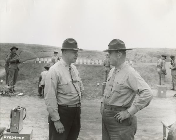 Two Marine captains talking at a Marine Corps rifle range. In the background, Marines are shooting at targets on a far hill.