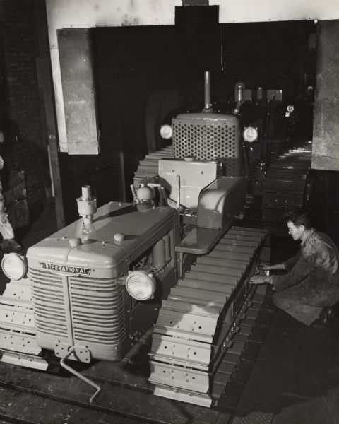 "Freshly painted" crawler tractors (TracTracTors) "emerge from 190 ft. gas-fired drying oven" at International Harvester's Tractor Works. A factory worker is crouched down near the side of a tractor.