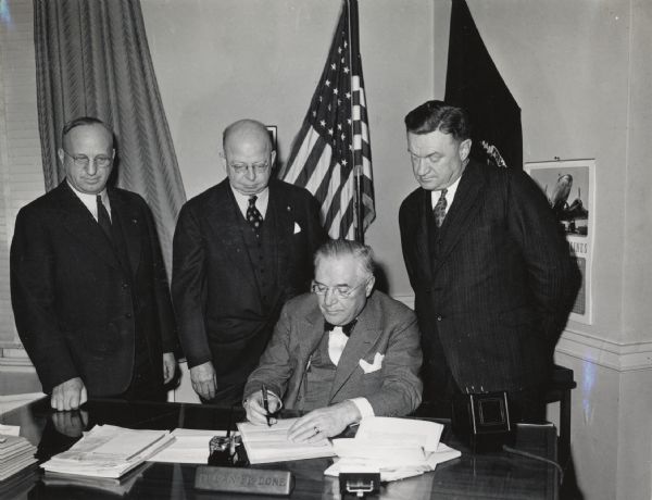 Governor Julius P. Heil sits at a desk surrounded by members of the Wisconsin Salvage Committee.
