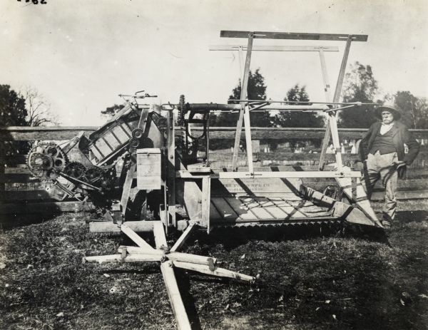 Man standing outdoors beside a Plano grain binder. On the machine is stamped: "Manufactured by The Plano Mfg. Co. / Plano Ill's." There appears to be a cemetery with trees in the background.