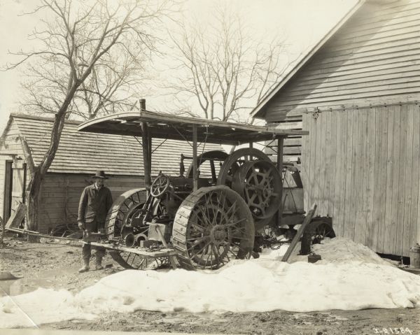 Man standing next to an International friction drive tractor, parked in the snow next to a farm building.