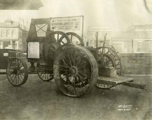 Tractor parked outdoors on what appears to be a loading dock. A sign on the tractor reads: "Old No. 13, International Harvester Co., Sold in 1906 - used for 16 yrs. Still going Strong . . ." A sign on the left reads: "See Me At The Wheat Show."