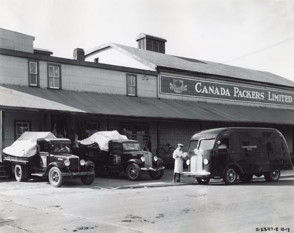 Men working outside the Canada Packers Limited Warehouse. There are three trucks, including an International Metro parked in front of the warehouse near the loading dock.