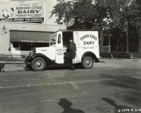 View from across street of a man holding a carton of milk bottles and standing next to an International truck owned by Border Cities Dairy. The truck is parked in front of the Border Cities dairy building.
