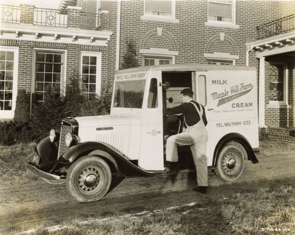 A man gets into an International truck owned by Maple Hill Farm Dairy. The truck dairy truck which is parked on a dirt driveway in front of a brick building. The photographer's shadow is visible in the foreground.