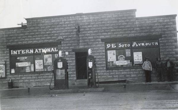 Exterior view of the Fleming Motor Company, an International Harvester dealership. Three men are standing on the right, and a gas pump is in front of the entrance. Signs on the building advertise "Plymouth" and "DeSoto".