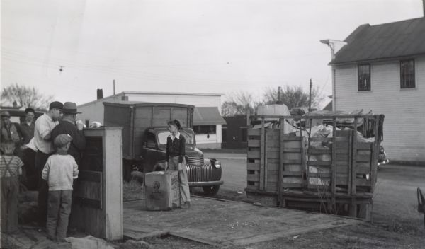 A group of men and boys are outdoors weighing metal collected in a wartime scrap drive. In the background is a truck and industrial buildings.