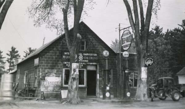 Exterior of the Sperstad Hardware and Implement Company, an International Harvester dealership. Two Deep Rock Gasoline pumps are near the front entrance. Two trees have posters for "Salvage for Victory." The dealership was involved in a statewide scrap drive effort.