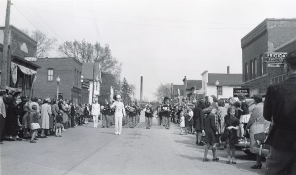 A high school band marches down the street during MacArthur Week, a state sponsored scrap drive effort. Crowds of children and adults line both sides of the street.