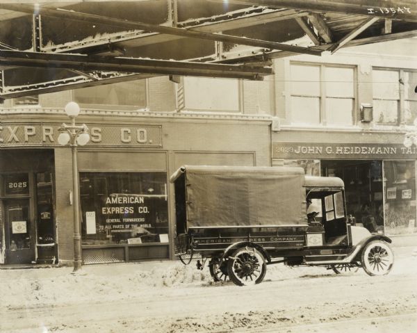 International Model F, or 31 truck owned by the American Express Company parked near a snowy curb outside the company office. A man wearing a hat is in the driver's seat. Above the street is a bridge or elevated railroad.