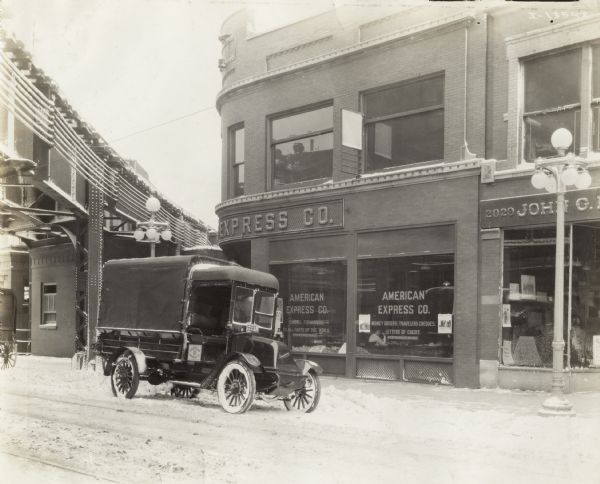 International Model F (or 31) truck owned by the American Express Company. The truck is parked in the snow outside the company office. A man wearing a hat is sitting in the driver's seat. Inside the office an employee is visible through the front window which advertises the company's services. Above the street on the left is a bridge or elevated railroad.