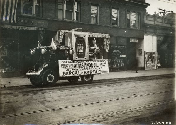 Highly decorated, perhaps for a parade, a International Model E truck owned by Barcal & Barta is parked outside the company storefront. A sign on the side of the truck advertises Atlas Floor Oil.