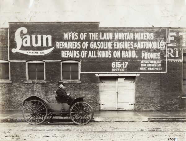 Two men wearing bowler hats are sitting in an International Model M  truck parked outside Laun Machine Co. A sign for the company is painted on the brick wall. The company manufactures mortar mixers, repairs gasoline engines and automobiles, and also does repairs.