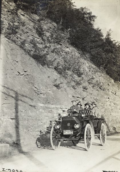 Man driving three women on scenic side road in an International model M truck. A steep hill, which has been cut away for the road, rises above them. The truck has a dealer license plate mounted on the front.