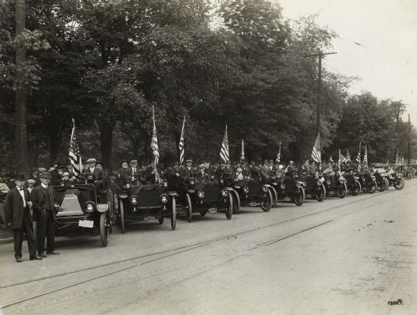 Large group of men and children posing in International trucks parked in a row on the side of a road. An American flag is held by one of the men in every truck. Some of the trucks may be Model E trucks.