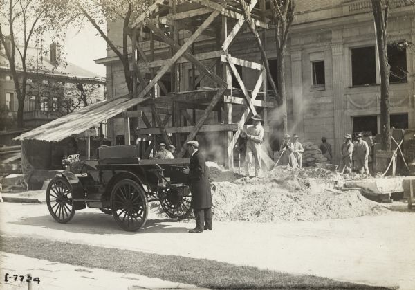 A man is standing at the back of an International truck holding a small chest or case. Behind him a group of construction workers are working near the scaffolding at the side of a building. The truck may be a Model M.