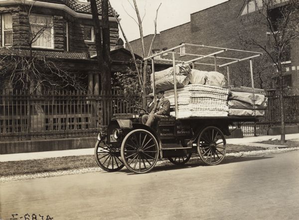 Two men on an International truck operated by Rhein Bros., mattress company. The truck is parked outside a residence and is loaded with  mattresses, box springs, and other bedding materials.