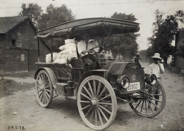 Man seated in an International model M truck loaded with sacks of flour, and with letters along the roof that read: "Crescent Roller Mills." Beside the truck is a young boy (possibly son?) barefoot, and wearing a hat. In the background on the left is a barn or shed.