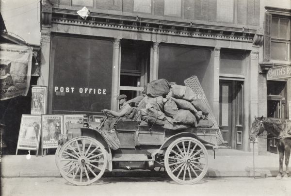 Man in the driver's seat of an International model M truck parked along a sidewalk outside a post office. Loaded on the truck are sacks and wire fencing. A horse is standing behind the truck on the street. Along the sidewalk outside the post office are movie posters for "The Godfather," "The Rival Barbers," and other films.