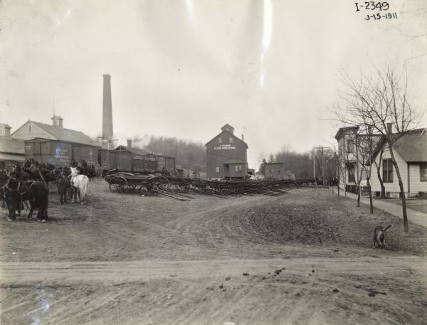 Over 15 manure spreaders near railroad cars in small town. They may have been recently transported from Chicago via the Chicago and Northwestern railroad company. Also pictured is a group of horses used to pull the spreaders.