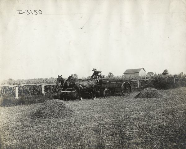Man and boy (possibly father and son) on farmland near a cornfield operating a horse-drawn No. 3 Clover Leaf manure spreader. A barn is in the background.