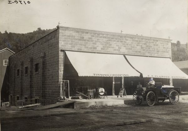 Man parked in an International Model M truck outside the storefront of E.W. Plaisted Hardware. Two men are seated just outside the store along with a small "20th Century" wagon, walking plow, a washing machine and other items. Advertised on the storefront are oils, greases, tinning, roofing and Eisenlohr's Cinco cigars.