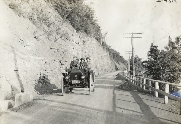 Man driving three women in an International Auto Wagon(?) on scenic unpaved road. The truck has a dealer license plate mounted on the front. A steep hill, which has been cut away for the road, rises above them on the left, and a guard fence is on the right side of the road.