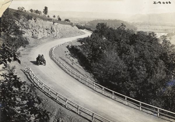 Elevated view of a man driving three women in an International Auto Wagon(?) along a scenic unpaved road overlooking a valley. There is a guard fence on both sides of the road.