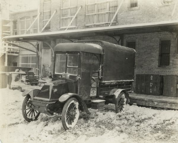 International model F, or 31 truck used by American Express Company for deliveries. The truck is parked in snow, possibly outside of the delivery depot of American Express. On the depot's platform are American Express crates for shipment. The truck is insulated for cold weather.