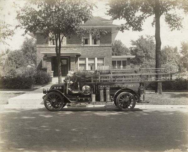 View from across the street of an International Model F or 31 truck used as a fire engine. The truck is parked outside a suburban residence. A young girl stands on the front porch of the home.