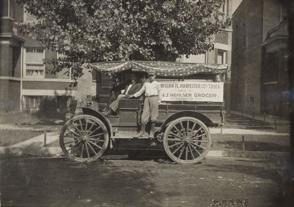 Man in driver's seat and a youth on the running board of a parked Internaitonal Model M truck used by H.J. Nehlsen Grocer. A sign on the side of the truck states that the truck is an International Harvester Company truck used by H.J. Nehlsen Grocer.