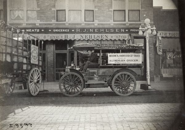 View from across the street of a man in the driver's seat of an International Model M truck used by H.J. Nehlson Grocer. The truck is parked in front of the Nehlson store front, is located at 4708 N. Kedzie Avenue. A large motor truck on the left with high open side gates is loaded with sacks.