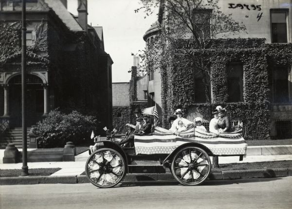 Man in driver's seat, with women and several small children (some or all possibly family?) in an International auto wagon parked near a curb on a residential street. The auto wagon is decorated with American flags and other patriotic banners.