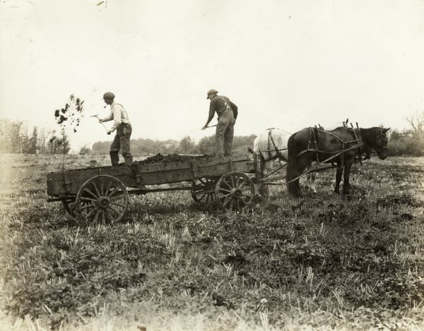 Two men with shovels on a horse-drawn manure spreader.