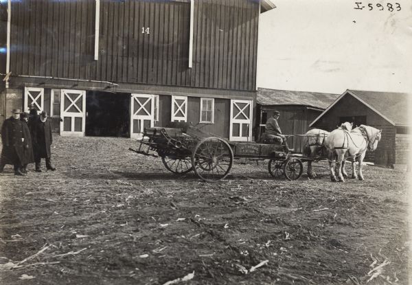 Man in a farmyard outside a barn on a Kemp Low Steel manure spreader pulled by two horses. On the left are a group of men wearing overcoats and hats.