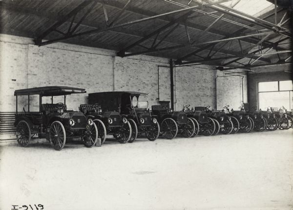 Approximately ten different models of motor trucks stored inside of a warehouse, possibly at a factory or branch house.