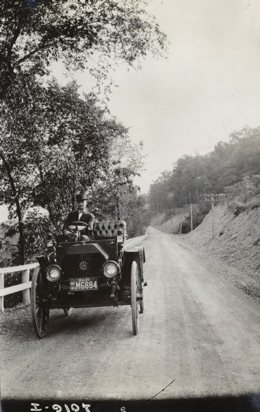 Well-dressed man driving an International Model M truck along a rural dirt road lined with trees. There is a guard fence on the left side of the road.